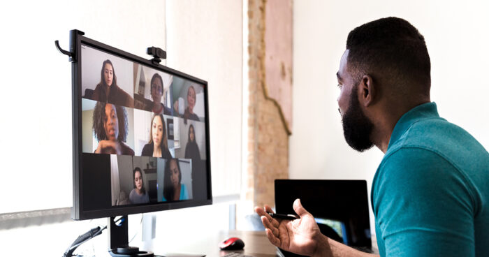 An adult man gestures while talking with a group of colleagues during a virtual staff meeting via video conference.